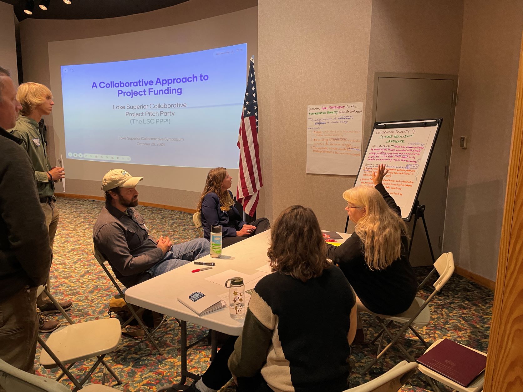 Participants at the 2024 LSC Pitch Party working in small groups, with a presenter at the front and conservation priority notes on a whiteboard