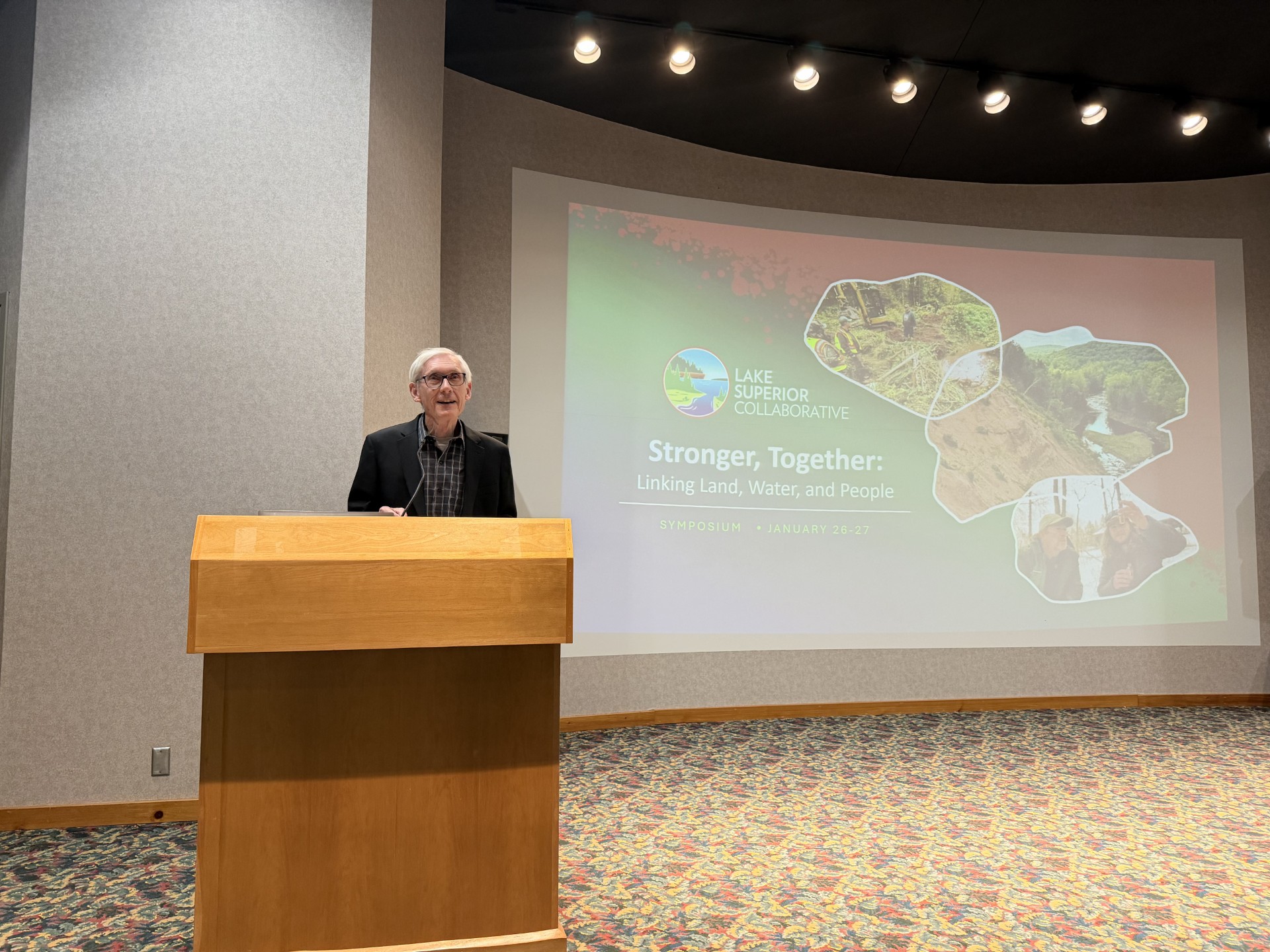 Governor Tony Evers addressing the 2026 LSC Symposium group at a podium with the Stronger Together symposium graphic projected behind him